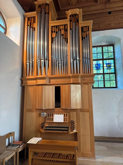 This is a full-frame, eye-level, interior shot of a pipe organ in the church of Bévilard, Switzerland. The organ is made of light-colored wood and features rows of polished metal pipes. The keyboard and controls are visible at the front, with sheet music placed on a stand above the keys. To the right of the organ, a stained-glass window depicts two heraldic shields, one with a bird and the other with a rooster, against a green background of trees. A circular window is visible on the left side of the image. The walls are white, and the ceiling is made of dark wood planks. In the foreground on the left, a wooden chair with a cushion holds a stack of notebooks and books. The floor is tiled with light and dark squares, and a dark metal grate runs along the base of the wall. The scene is well-lit, with natural light coming from the windows.
