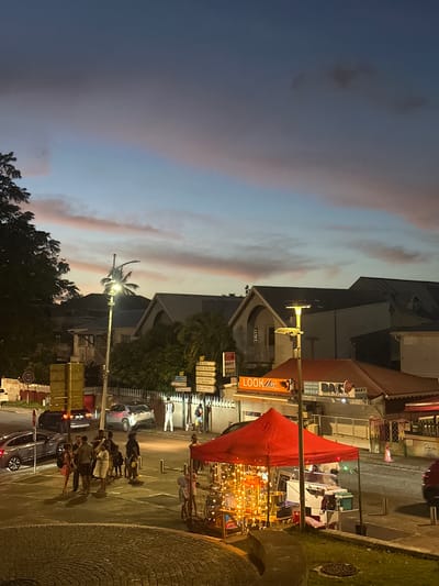 A nighttime street scene in Le Gosier, France, showcases several key elements. Under a twilight sky, a street corner is lit by streetlights. A group of people gathers, their silhouettes defined against the illuminated pavement. Cars parked along the road suggest a public space. A street vendor operates under a red awning, decorated with strings of lights. Buildings line the street, storefronts visible with their signs, including one for "Look Hair." Street signs in French direct traffic. The scene combines pedestrian activity and urban elements in the evening atmosphere of Le Gosier.