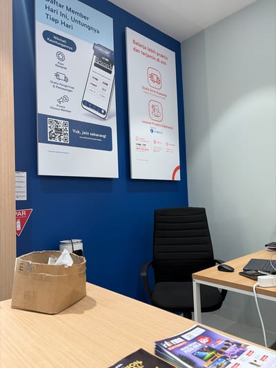 The image shows a retail store interior, likely an electronics store, with promotional posters on a blue wall. A wooden desk with a laptop, mouse, and some cables is visible, along with a black office chair. To the right, a stack of magazines or flyers with promotional offers, including "PESTA BOLA 50% DISKON SPESIAL," is displayed. A cardboard box with some paper inside sits on the desk.

Prominently featured are two large posters. The first poster, on the left, announces "Daftar Member Hari Ini, Untungnya Tiap Hari" (Register as a Member Today, Get Benefits Every Day) and highlights benefits like "Nikmati Keuntungannya" (Enjoy the Benefits), "Poin Rewards," "Gratis Pengiriman & Pemasangan" (Free Delivery & Installation), and "Promo Khusus Member" (Special Member Promotion). It also shows a smartphone displaying an app with various brand logos.

The second poster, to the right of the first, states "Belanja lebih praktis dan terjamin di sini" (Shop more practically and with assurance here) and advertises "Layanan Proteksi Elektronik" (Electronic Protection Service) powered by ZURICH. It also includes information about free delivery and installation.

Visible text includes brand names like "informa," "ZURICH," "THYS," "KELS," and "EYESOUL," along with a QR code and a website address "bit.ly/REGISTRASIINFORMA." There is also contact information for a "Chat Center" and a phone number. The overall scene suggests a retail environment focused on customer acquisition and promoting product benefits and services. The lighting suggests it's daytime inside a well-lit store.