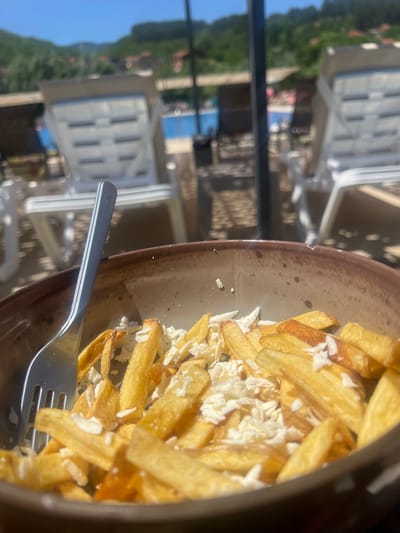 A close-up, shallow depth of field shot shows a bowl of french fries with crumbled white cheese, garnished with a fork, on a table with a swimming pool and sun loungers in the background. The location is outdoors with a clear blue sky and lush green hills in the distance. The scene appears to be a resort or restaurant setting, likely in the city of Delchevo, North Macedonia, given the context provided. The focus is on the food, highlighting its texture and presentation.