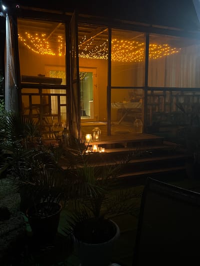 The image depicts the exterior of a screened-in porch or gazebo at night in Serhiivka, Ukraine. The structure is illuminated by strings of warm-toned fairy lights draped across the ceiling. Inside, a table is set, suggesting a dining or gathering space. Several glass lanterns with lit candles are placed on the wooden steps leading up to the porch, casting a soft glow. Potted plants, including palm-like foliage, are situated in the foreground. The overall atmosphere is cozy and inviting, despite the darkness. The presence of the fairy lights and candles creates a festive or romantic ambiance.