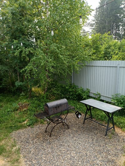 The image depicts a backyard scene in Kurlek, Russia, featuring a rustic barbecue setup. A dark-colored metal barbecue grill, with intricate scrollwork on its legs, sits on a gravel path. To the right of the grill is a simple, dark metal table with a rectangular top. Surrounding the gravel area is a patch of green grass, and beyond that, lush vegetation and trees. A light-colored metal fence forms the backdrop of the yard. String lights with clear bulbs are visible hanging above the scene, suggesting an outdoor gathering space. The overall atmosphere is one of a serene, natural outdoor environment.