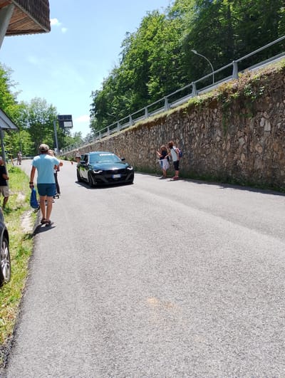 In the city of Rieti, Italy, on a sunny day, a black BMW car is driving on an asphalt road bordered by a stone wall and lush green trees. On the left side of the road, a man wearing a blue t-shirt and shorts walks away from the camera, carrying a blue plastic bag. Several other people are visible along the roadside, some walking and others standing. Two individuals, a woman and a child, are standing near the stone wall, looking towards the car. The car has its headlights on and a license plate that appears to read "DC428MX". The road curves to the right and slopes slightly uphill. The sky is blue with a few scattered clouds.