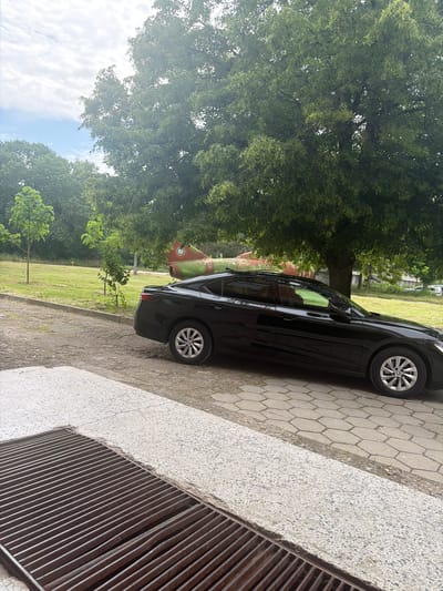 A black car is parked on a paved area adjacent to a grassy field with trees. In the background, there is an old red and green airplane on display. A metal grate is visible in the foreground. The location is Dolna Mitropolia, Bulgaria.