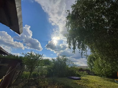 This image presents a vibrant, sunlit outdoor scene, characteristic of a garden or backyard in Polanki, Russia. The upper half is dominated by a bright blue sky filled with picturesque white cumulus clouds. The sun, shining brightly from behind a cloud in the upper middle, creates striking crepuscular rays that extend across the sky.
In the foreground, a dark, angular structure, possibly a roof overhang or part of a building, is visible in the upper left corner. The right side of the frame is filled with the dense green foliage of a large, leafy tree, likely a birch, whose branches extend from the top and right.
The midground features lush greenery. On the left, smaller trees and bushes stand near what appears to be a rustic wooden structure or fence. The ground below transitions from a patch of cultivated earth with emerging green shoots to a well-maintained grassy area. A dark purple inflatable object, possibly a mattress or a pool float, lies on the grass in the mid-right.
In the background, more trees and the subtle outlines of distant building roofs can be seen, suggesting a rural or semi-rural setting. The overall atmosphere is one of natural beauty and peacefulness. The bright sun and scattered clouds indicate a clear and pleasant day, likely late morning or early afternoon. There is no visible text in the image.