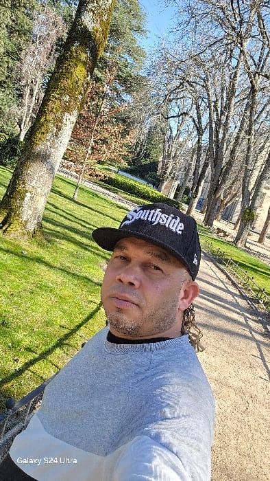 An adult male is captured in a selfie within the formal gardens of the Royal Palace of La Granja de San Ildefonso, Real Sitio de San Ildefonso, Spain. The subject, positioned centrally in the foreground, wears a black baseball cap with "Southside" embroidered in white, a grey and white two-tone long-sleeve shirt, and has a short beard and curly hair visible at the back of his head. He looks directly at the camera. To his left, a large tree trunk covered in green moss or lichen is visible. The ground consists of bright green grass to his left and a light-colored gravel path, bordered by a low dark fence, extending into the background on his right. Numerous bare deciduous trees line the path and frame the scene, suggesting winter or early spring. In the far background, architectural elements of the palace, including stone buildings with arched features, are partially visible through the trees, alongside what appears to be a body of water. The sky is clear blue, and bright sunlight casts distinct shadows across the landscape. The image contains a "Galaxy S24 Ultra" watermark in the lower-left corner.