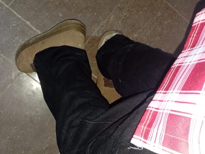 The image shows a close-up, overhead view of a person's legs and feet. The person is wearing dark wash jeans and tan, ankle-high boots. A red and white checkered fabric, likely a shirt or blanket, is draped over the right leg. The scene is set on a floor tiled with light brown and grey square tiles. The lighting suggests it may be indoors or during a cloudy day, as there are no distinct shadows indicating bright sunlight. The setting appears to be casual. There is no discernible text in the image.