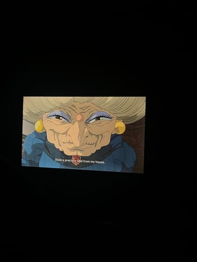 The image is of an animated character on a screen, possibly a television or computer monitor. The character is an elderly woman with a large, wrinkled face, pale skin, and light blue eyes with purple eyeliner. She has a large nose and is wearing yellow earrings. Her hair is white and styled in a curled updo. The character is wearing a blue garment with dark blue shading. Text near the character says "Stole a precious seal from my house." The background appears to be a dark wood with swirling patterns. The image is set against a completely black background. There are no other details that would suggest a location. Therefore, determining its location in Tepoztlán, Mexico, is impossible.