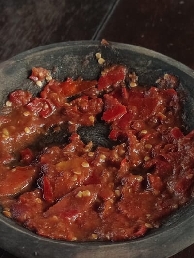 A close-up image shows a dark gray stone mortar filled with red chili paste, commonly known as sambal, in Kecamatan Beji, Indonesia, a location at latitude -7.605, longitude 112.759. The paste is composed of finely crushed red chili peppers and other unidentified ingredients. It appears to be wet and freshly made, with some larger pieces of chili pepper visible in the mixture. The mortar appears to be made of rough stone and has a dark gray, worn appearance. The texture of the sambal suggests it is a spicy condiment commonly used in Indonesian cuisine.