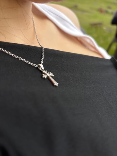 A close-up shot depicts a person wearing a black garment with a silver cross pendant necklace. A white undergarment is also visible. The background is blurred and out of focus, showing green hues suggesting foliage or grass. There is no indication of the location being Tlik, Armenia.