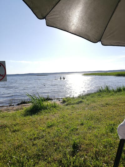The image captures a serene daytime scene at a lakeside, likely in the Перабродскі сельскі Савет region of Belarus. The sun is high in the sky, casting a bright glare on the water's surface and suggesting a warm, clear day. The foreground is dominated by lush green grass, with some sandy patches near the water's edge and reeds growing along the shoreline.
In the middle ground, several people are visible in the lake, appearing to be swimming or enjoying the water. A lone duck is also present on the water. In the far distance, a line of trees marks the horizon. The top portion of the image is partially obscured by the shadow of a large umbrella, indicating that someone is likely enjoying the shade while observing the lake. To the left, a sign with a red circle and a diagonal line over a silhouette suggests a prohibition, possibly related to water activities. The overall mood is peaceful and relaxed, evoking a sense of summer leisure.