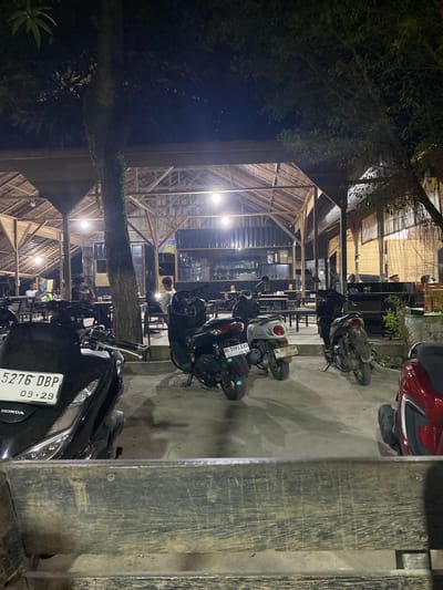 The image captures an evening scene at an outdoor or semi-outdoor establishment, likely a cafe or restaurant, in Seuneubok Teungoh, Indonesia. The foreground and mid-ground are occupied by numerous parked motorcycles and scooters on a concrete surface. In the immediate foreground, a sturdy wooden barrier partially obscures a black Honda scooter with license plate '5276 DBP', showing an expiry date of '09-29'. Other visible scooters include a white one with license plate 'BL 8391 ZAS' (expiry '12-25') and another white one displaying 'BL 4117 OBP' (expiry '06/25'). A red scooter is also visible on the far left.
The main structure, brightly lit by artificial hanging lamps, features a distinctive wooden frame and what appears to be a thatched or bamboo roof, providing a rustic aesthetic. Inside, several individuals are visible, seated or standing, suggesting patrons socializing or dining. A tall tree trunk ascends through the middle of the frame, with dense green foliage from surrounding trees prominent on the left side and upper portions of the image, emphasizing the outdoor setting. The presence of 'BL' on the license plates further situates the scene in Aceh province, Indonesia. The overall atmosphere suggests a lively night spot.