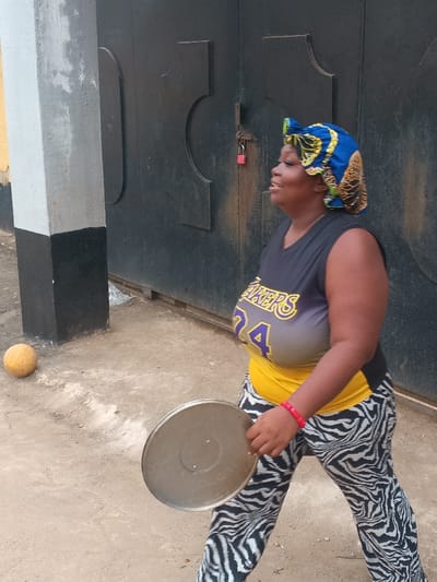 An adult female is standing outdoors on a light brown dirt surface in Itu, Nigeria. She faces towards the viewer's left, her mouth slightly open. She wears a blue, yellow, and green patterned fabric headwrap. Her attire includes a black and grey sleeveless v-neck top with "LAKERS 24" in yellow and purple lettering, a yellow garment visible beneath the top, and black and white zebra-print pants. A red beaded bracelet is on her right wrist. She holds a circular, metallic pan lid in her left hand, angled downwards. To her left, a light yellow spherical object rests on the ground beside a grey concrete pillar with a black base. Behind her is a dark-colored gate featuring raised decorative panels and secured by a visible red padlock.