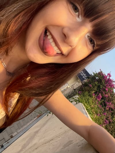 The image depicts a close-up, tilted selfie of a young female positioned in an outdoor urban setting in Kefar Sava, Israel. The subject's face, predominantly illuminated by warm, natural light, occupies the left and central areas of the frame. She presents with an open-mouthed smile, exposing white teeth and an extended tongue. Her eyes are light brown, and the lower lash line exhibits a light-colored cosmetic application. Her hair is long, brown, with visible reddish highlights, and styled with bangs. A silver-colored chain necklace with a small pendant, possibly displaying Hebrew text such as "אמא" (Ima, meaning Mother), is worn around her neck, partially obscuring a light-colored garment. Her right forearm rests horizontally on a light-colored concrete or stone railing in the lower right foreground. In the background, to the right of the subject, a dense bush with vibrant pink-purple bougainvillea flowers is visible. Above the bougainvillea, a dark blue sign with white Hebrew text, clearly displaying "חניה" (ḥanaya, meaning parking), is mounted on an indistinct building structure, indicating a parking area within the city. The sky above is a clear light blue.