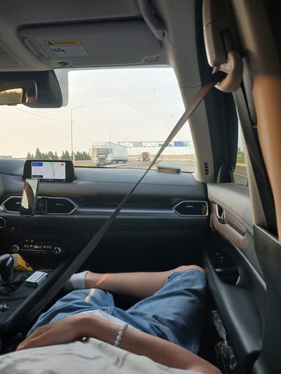 The image is taken from the interior of a car looking forward, showing the dashboard and the view through the windshield. A person's legs, wearing denim shorts and white socks, are visible in the foreground, resting on the passenger seat. A seatbelt is diagonally stretched across the frame, connecting to a buckle on the right side. The dashboard features a GPS navigation screen displaying a map and route, a smartphone mounted on a holder, and car climate controls. Outside the car, a highway is visible with other vehicles, including trucks and cars, and trees line the road. The sky is overcast with some light. The scene appears to be a daytime road trip.