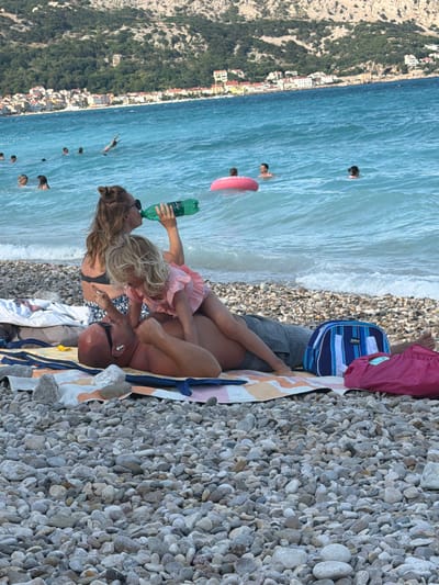 This is a vertical, eye-level shot of a pebbly beach with a clear blue ocean and a green, tree-covered hillside in the background, with a town nestled on its slope. The scene is bathed in bright sunlight, suggesting a warm day.
In the foreground, two people are lying on a beach towel. A woman with blonde hair, wearing a pink ruffled top and a black bikini, is drinking from a green plastic bottle. She is looking at a man who is lying next to her, with his head turned towards her. He is wearing sunglasses and appears to be bald. His body is partially covered by the woman's legs. A blue and white striped backpack and a pink bag are placed behind them.
Further out in the water, several people are visible swimming or wading. One person is on a pink inflatable ring. Another person appears to be in mid-dive. The water is calm with small waves gently lapping the shore.
The hillside in the background features a dense green forest and a scattering of white buildings, characteristic of a coastal town. The pebbles on the beach are predominantly grey and white, of various sizes. The overall mood of the image is relaxed and summery.