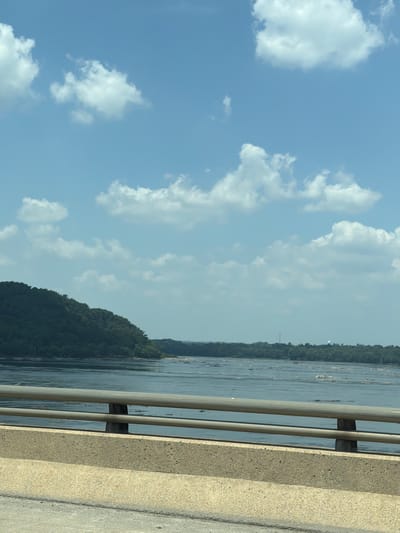 The image shows a wide river with choppy water, dotted with rocks. The river is bordered by a dense, tree-covered hill on the left and a flatter, tree-lined shore on the right. A bright blue sky with scattered white clouds stretches above. In the foreground, there is a concrete barrier and metal railings of a bridge, suggesting the photo was taken from a vehicle moving across it. In the distance on the right shore, two structures are visible: a tall, slender tower and a white, spherical water tower. This scene depicts a natural landscape from a vantage point on a bridge.