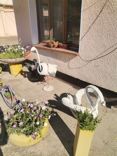This is an outdoor scene in Dekov, Bulgaria, featuring garden decorations made from repurposed tires. In the foreground, a bright yellow tire planter overflows with pink and white petunias. To the right, a white swan sculpture, also crafted from tires, is depicted in a coiled position with its head raised. Behind the swan, a tall, pale yellow rectangular planter contains green foliage. Further back and to the left, a white stork sculpture with a red beak stands on thin red legs, positioned near a larger, gray tire planter holding more pink and white flowers. The ground is paved with concrete, and sunlight casts shadows. A textured, cream-colored wall with a window is visible in the background, with electrical wires running along its surface.