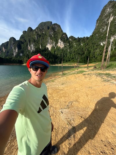 A man stands on a dry, cracked, sandy shore, holding a camera to take a selfie. He is wearing a light green t-shirt with a black Adidas logo on the chest, sunglasses, and a red hard hat over a blue cap. Behind him, a turquoise lake is visible, with towering, verdant limestone cliffs in the distance. The sky is clear and blue. Several dead trees stand in the shallow water and on the shore, their branches stark against the lush green of the cliffs. The scene is consistent with the landscape of Khao Phang, Thailand, known for its limestone karst formations and reservoirs. The man's shadow is cast long on the ground to his right.
