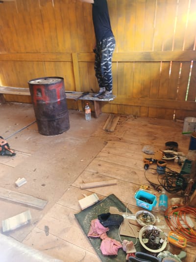 The image depicts an interior scene, likely a workshop or storage area, with a focus on construction or repair activities. A person, partially visible from the waist down, is standing on a wooden platform against a yellow-painted wooden wall. They are wearing camouflage-patterned pants and dark shoes.
In the foreground, a large, rusty metal barrel with red markings is prominent. Scattered across the wooden floor are various tools and materials. These include a chainsaw, a power drill, a blue plastic container filled with screws, a white bucket, electrical cords, and several pieces of wood. There are also two bowls containing what appear to be nails and other small metal objects, and a pink cloth lies near a black shoe. The overall environment is rough and appears to be in a state of disarray, consistent with a work in progress. The lighting suggests it is daytime, with natural light entering the space from the right side of the frame. The location is specified as Islambaevo, Russia.