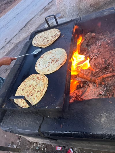 Three pieces of flatbread are being cooked on a large, flat metal surface above an open fire. The fire is burning brightly with yellow and orange flames. A hand holding a spatula is visible, presumably turning the bread. The cooking surface is black and shows signs of wear. The flatbreads are golden-brown and have a slightly uneven surface. The cooking apparatus appears to be a rudimentary, outdoor oven.
