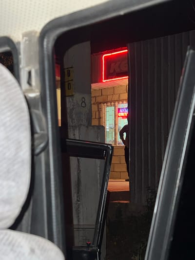 The image, taken at night, shows a view through the open door of a vehicle looking towards a building with a corrugated metal exterior. In the background, a neon sign glows red with the letters "KG" visible. A person is partially visible inside the building through a window, illuminated by internal lights and the neon sign. The building appears to be constructed of cinder blocks. To the left of the building, a column has the number "8" painted on it. The overall scene suggests an urban or semi-urban setting at night, potentially a commercial establishment. The location is identified as Mukur, Kyrgyzstan.