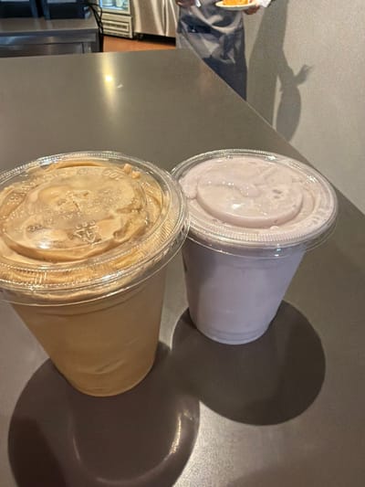 The image depicts an indoor scene within a commercial establishment, specifically in Fraccionamiento Los Heroes Queretaro, Mexico. Two plastic cups with domed lids containing beverages are positioned on a dark gray countertop in the foreground. The left cup holds a light brown or caramel-colored liquid, while the right cup contains a light purple liquid. Both lids display embossed text, including "100% RECICLABLE" and "PET". Strong, distinct shadows from both cups are cast on the countertop, extending towards the lower right of the frame. In the blurred background, behind the counter, a person is partially visible from the chest down, wearing a dark apron or uniform. This individual is holding a small white plate with a yellow-orange item on it, possibly a pastry or cake slice. Stainless steel equipment, likely refrigeration units or service counters, is also visible behind the person. A pale wall forms the backdrop.