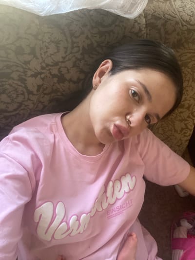 A young woman with dark hair, wearing a pink t-shirt with white lettering, is lying on a patterned sofa. She is looking directly at the camera with her lips pursed. She has a nose ring and earrings. A small child's foot is visible in the lower right portion of the image. The background appears to be an interior space. The city of Romny, Ukraine is not mentioned or depicted in the image.