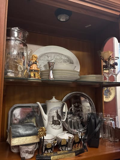 A china cabinet features plates with deer motifs, stacked and held by a silver stand. A Hummel figurine sits below. A silver tray is on the right side of a glass shelf, along with glasses, a small black purse, and a deer-themed tea set. Decorative wine glasses and a commemorative plate are visible on the top shelf. The cabinet is made of wood.