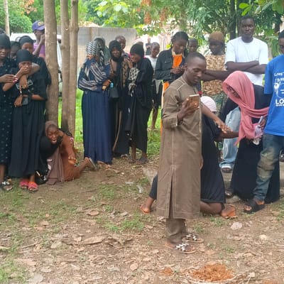 The image depicts a group of people gathered outdoors, possibly in Andaha, Nigeria. Most of the individuals appear to be in mourning, with several women visibly upset and being comforted by others. The mourners are dressed in varying shades of black and dark-colored clothing. One woman is on the ground, being held back by another person. A man in a brown robe is standing and looking at his cell phone. The environment appears to be a grassy or dirt area with trees and a building in the background. There is a small mound of dirt in the foreground.