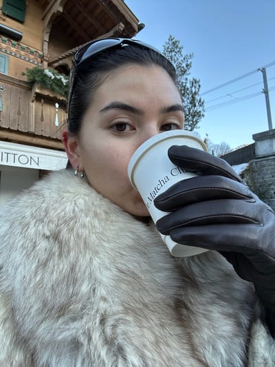 A woman is depicted outdoors in Gstaad, Switzerland, drinking from a white paper cup. She is positioned facing forward, with her right hand, encased in a dark brown leather glove, holding the cup to her lips. Her dark hair is pulled back, and dark-lensed sunglasses rest on her head. She wears small, silver-toned hoop earrings. A light-colored fur coat covers her upper body. The paper cup bears the visible text "The Matcha Club." In the background to her left, a traditional wooden chalet-style building with intricate carvings, a balcony, and green shutters is visible. Below the balcony, the illuminated letters "VUITTON" are apparent, indicating a Louis Vuitton store. Evergreen foliage with traces of snow adorns the balcony railing. To her right, overhead electric wires are visible against a clear blue sky, above a grey stone structure, possibly a bridge or retaining wall. A coniferous tree is also present in the right background.
