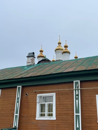 The image shows an exterior view of a wooden building with a green metal roof in Kstining, Russia. The building's facade is constructed from horizontal brown wooden planks, adorned with vertical green and white decorative elements at the corners. A white-framed window with visible internal shutters is centered on the visible portion of the wall. Above the building, the golden domes of several Orthodox churches are visible against a pale, overcast sky. A brick chimney with a metal cap protrudes from the green roof. The scene is overcast and appears to be a daytime shot. There are no people or animals present in the image.