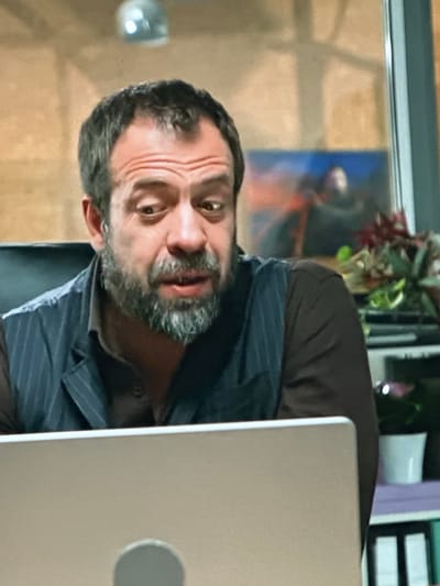 A man with short, dark hair and a beard is looking to his left with a surprised expression. He is wearing a dark vest over a brown shirt. A laptop is in front of him, partially obscuring his lower face. In the background, there is a shelf with plants and binders, and a blurred image on the wall. The overall lighting suggests an indoor setting, possibly an office. The scene is set in the city of Dondușeni, Moldova.