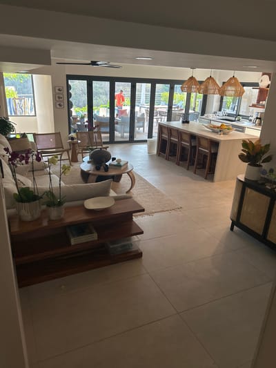 The image shows the interior of a house in Playa Herradura, Costa Rica, located at latitude 9.658 and longitude -84.673. The scene is a living room with a couch, a coffee table, and a large open space with a kitchen visible in the background. The couch is white and has pillows and blankets. There are two potted plants on the couch, both with white pots. The coffee table is made of wood and has a black vase and a book on top. The kitchen has a counter with a white countertop and a sink. There are bar stools around the counter. There are three woven pendant lights hanging over the counter. The kitchen floor is beige tile. There is a large white wall in the background. The living room floor is a mix of beige tile and a large area rug. There is a set of large sliding glass doors leading outside, and the area outside is visible through the doors, with a pool and a patio with chairs. There is a person wearing red standing outside on the patio. The living room is sparsely furnished and decorated, with a minimalist style. The scene is bright and well-lit, and the light appears to be coming from natural light sources.