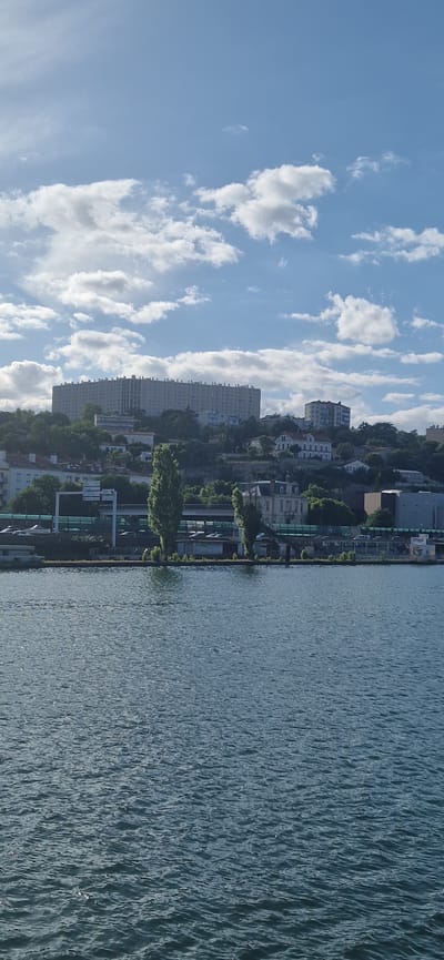 The image presents a panoramic view of the Saône River and the hillside of La Mulatière, France, under a blue sky scattered with white cumulus clouds. The lower half of the frame is occupied by the river's surface, which is dark blue-green and features numerous ripples creating bright specular reflections of sunlight.
The opposite riverbank, from left to right, shows a narrow strip of land containing several tall, slender deciduous trees. Behind these trees and along the bank, a multi-lane road or highway is partially visible, protected by green sound barriers. Traffic signs are visible above the road on the left. Structures along the immediate bank include industrial-style buildings, one featuring discernible graffiti text.
Above the road, the hillside ascends, densely covered with trees and various residential buildings. Prominently positioned on the upper left of the hill is a large, elongated, light-colored multi-story apartment complex, characterized by numerous windows and a distinct architectural pattern along its roofline. To the right of this dominant structure, other multi-story residential buildings and detached houses are interspersed with lush green vegetation across the remainder of the hillside. No individuals or discernible human activity are visible within the frame.