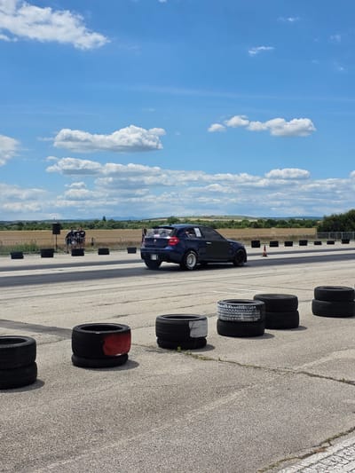 An outdoor, eye-level, medium shot captures a blue BMW racing car on a paved track under a bright, partly cloudy blue sky. The car, with a distinctive rear spoiler and racing decals, is positioned in the right half of the frame, angled slightly away from the camera. In the foreground, several stacks of used tires, some painted with red and white markings, are scattered across the track.

In the mid-ground, a group of three individuals are visible standing near a speaker on a stand, with a metal fence separating them from the track. Beyond the fence, a dry, agricultural landscape stretches towards a distant, hazy horizon, suggesting a rural or semi-rural setting. The presence of tire barriers and a racing car indicates a track event or practice session. The image appears to be taken at the Drag Racing Track Akatsievo, located near Akatsievo, Bulgaria, given the context of the racing car and track setup. The lighting suggests it is daytime, likely late morning or early afternoon.