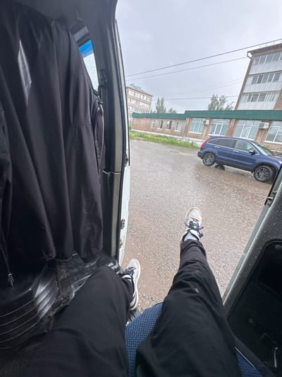 This image presents a first-person perspective from inside a vehicle, looking out onto a wet street in Mozhga, Russia, under an overcast sky. The overall scene is oriented as if rotated clockwise.

In the foreground, a person's legs and feet are visible. The person is wearing black pants, which have some light smudges, and light-colored sneakers with dark laces and white soles. One foot is positioned outside the vehicle, near the open door, resting on a textured, black surface. The interior of the vehicle shows part of a blue patterned seat on the right and a large black fabric item, possibly a folded jacket or bag, occupying the left side of the lower frame.

Outside the vehicle, the asphalt or concrete road surface appears wet and speckled, indicative of recent rain. A dark blue SUV or crossover vehicle with grey wheels is parked on the left side of the street. In the mid-ground, against a grey sky, several multi-story brick buildings with numerous windows and visible air conditioning units line the street. Utility lines stretch across the sky in the distance, and glimpses of green trees are visible between the buildings.

The scene depicts a mundane, daytime moment in an urban or suburban setting, marked by wet weather. No specific activity is clearly shown other than the person's feet resting outside the vehicle. There is no visible text in the image.