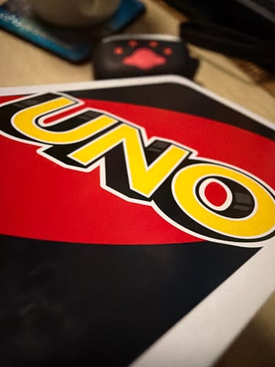 The image displays a close-up view of a Uno card, with the iconic "UNO" logo prominently featured. The card is predominantly red and black, with the yellow letters of the logo outlined in white. In the background, out of focus, are other objects, including what appears to be a small black pouch with red paw prints. The surface the card is resting on is textured and appears to be a table or similar flat surface. The lighting suggests an indoor setting. There is no visible indication of the specific location in Castillo Bajo-conejito, Spain.