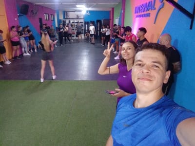 The image displays an indoor fitness facility located in Quilmes, Argentina, occupied by approximately 17-20 individuals. In the immediate foreground, a light-skinned male with short dark hair, wearing a blue short-sleeved athletic shirt with a white Nike logo, is positioned centrally, capturing a selfie. He exhibits a slight smile and faces the camera directly. Immediately behind and to his left, a light-skinned female with dark hair pulled back, wearing a purple sleeveless athletic top with a white Nike logo, smiles towards the camera while holding her right hand in a peace sign. She holds a smartphone with a distinct pink/purple case in her left hand.
The facility's flooring consists of a green artificial turf section in the foreground, transitioning to a darker, possibly rubberized, surface further back. The walls are painted in bright, angular geometric patterns utilizing magenta, orange, blue, and yellow hues. On the blue and orange section of the back wall, a prominent white sign reads "INTERVAL," accompanied by an athletic-themed logo and smaller text below it.
In the mid-ground and background, other adults and young adults are visible, dressed in various forms of athletic wear including t-shirts, shorts, and sneakers. A group on the left side leans against a wall with pink and yellow stripes, primarily engaged with their smartphones. A woman with long blonde hair, wearing a dark top and dark shorts, stands facing away from the camera near the transition between the green and dark floor sections. On the right, near the "INTERVAL" sign, a group stands conversing; one bald male with a beard and tattoos is partially visible. The area is uniformly illuminated by overhead artificial lighting.