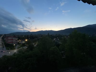 This image presents a panoramic view of the town of Zlatitsa, Bulgaria, captured during dusk or early night. The scene is dominated by a vast sky exhibiting a deep blue hue that gradually lightens towards the horizon, where a subtle band of orange-yellow light indicates the recently set sun. Clouds are scattered across the sky, with more prominent, darker formations on the left and lighter, wispy clouds towards the right.

In the foreground, dense, dark green foliage from several large trees obscures the lower part of the frame, suggesting the photo was taken from an elevated vantage point, possibly a balcony or a high window. A dark, indistinct structure, possibly a roof or awning, is partially visible in the top right corner.

Below the trees, the town is illuminated by numerous streetlights and lights from buildings, appearing as scattered pinpricks of light against the darkening landscape. On the left side of the image, a distinctive multi-story apartment building with alternating red and white horizontal stripes is clearly visible next to a road intersection featuring a marked crosswalk. Several cars are present on the roads, some with their headlights on.

Beyond the town and stretching across the midground, the dark silhouettes of mountains rise majestically, forming a dramatic backdrop against the twilight sky. The overall impression is one of peaceful transition from day to night in a mountainous, residential area. There is no visible text in the image.
