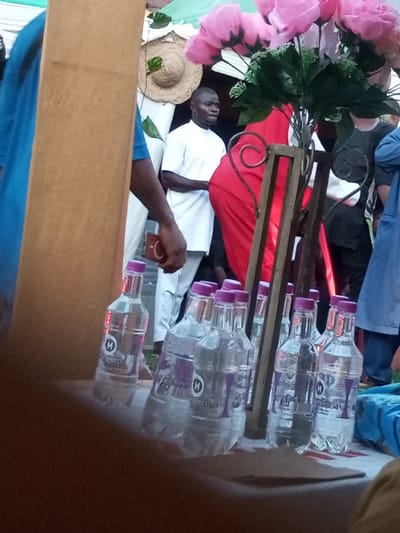 The scene in Amokvo, Nigeria, depicts a gathering, possibly an event, with multiple individuals and decorative elements. In the immediate foreground, a blurred, dark brown surface occupies the lower portion of the frame. On this surface, a collection of at least ten plastic water bottles, labeled "Mpero" with purple branding and purple screw caps, are arranged. To the left, a thick, light brown vertical column or pillar partially obstructs the view.
Behind the bottles and column, a dark metal stand with scrollwork, including heart-shaped motifs, supports a large artificial floral arrangement featuring multiple oversized pink roses and green foliage.
Several individuals are visible in the midground. A dark-skinned man with short dark hair stands prominently in the center-left, wearing a white tunic-style shirt with a collar and white trousers. His hands are clasped in front of him, and he gazes towards the right of the frame. To his left, a person wearing a vibrant red garment, possibly a wrapper or dress, is partially visible from the waist down, facing away. To the man's right, a person in a blue top is partially visible, with their right forearm and hand, holding a small brown rectangular object, visible near the water bottles. Further to the right, another individual wearing a blue garment is partially obscured.
In the background, a light-colored straw hat decorated with green leaves is suspended on a wall or structure. Overlapping fabric, appearing as light green and white canopy material, is visible at the top of the frame, suggesting an outdoor or semi-outdoor event setting. The overall lighting suggests daylight conditions.