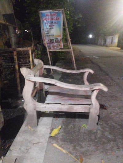 The image presents a dimly lit, quiet roadside scene at night, likely in a rural or semi-urban area of Kecamatan Jangkar, Indonesia. In the foreground, a rustic, light-colored wooden bench is positioned on a paved surface. The bench appears aged, with simple, handcrafted elements like curved armrests and back supports.
Dominating the midground is a large banner, supported by a wooden or bamboo-like frame. The banner prominently advertises "WARUNG GORENGAN" (fried food stall) with "Laros Manc" beneath it. Further text lists food and drink items: "ES CAMPUR" (mixed ice), "JAHU", "SOSIS" (sausage), and "TAHU WALIK". Small, indistinct pictures of various food items are visible at the bottom of the banner.
The background reveals a dark road or pathway receding into the distance, with faint light sources suggesting distant streetlights or buildings. Trees and foliage line the edges of the road and frame the scene. The time of day is clearly night, indicated by the overall darkness and artificial illumination. No people are visible, suggesting a deserted moment, perhaps between customers or after closing hours. A few fallen yellow leaves are scattered on the ground near the bench, and the concrete surface shows some cracks. The atmosphere is calm and still.