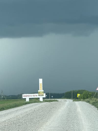 The image captures a rural road scene in Ильчигулово, Russia, under a somber, heavily overcast sky. Approximately two-thirds of the upper frame is dominated by dark grey to bluish-grey storm clouds, indicating inclement weather. A light-colored gravel road occupies the lower third, extending from the foreground and curving gently to the right as it ascends. The road surface is composed of small, light-toned stones, with discernible tire tracks in its center.
To the left of the road, a green grassy verge leads to a prominent white roadside monument. This structure features a central vertical pillar adorned with a golden stylized wheat stalk. Below this, a horizontal white sign displays the Cyrillic text "ДОБРОГО ПУТИ" (Dobrogo Puti), translating to "Have a good journey." Above the horizontal sign, a smaller rectangular sign contains additional text: "ХЭНЕРЛЕ ЮЛ!" and "СЧАСТЛИВОГО ПУТИ!", both conveying expressions of well-wishing for travelers, likely in Russian and a local language.
Further to the left, in the background, several utility poles with visible lines are interspersed across green fields, leading to a distant treeline. To the right of the road, a similar grassy verge is present. A yellow circular speed limit sign with "70" in black text is mounted on a pole, and a red and white triangular warning sign is partially visible towards the right edge of the frame. A dense line of green trees forms a forest that stretches across the mid-ground, following the contour of the road. No individuals or animals are present in the scene.