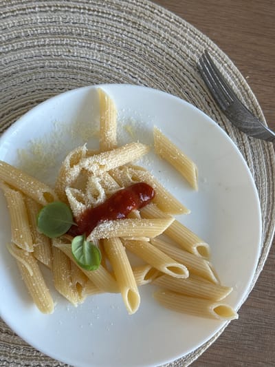 A white plate of penne pasta with grated cheese, a dollop of tomato sauce, and two basil leaves sits on a textured placemat. A fork is visible on the right side of the plate. The background is a wooden surface. This image is not located in the city of Гамово, Russia.