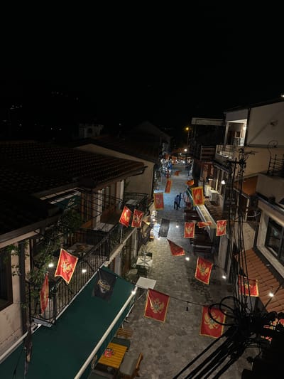 The image captures a narrow street in the city of Bartula, Montenegro, at night. The street is paved with cobblestones and lined with buildings that appear to be shops or restaurants, many with balconies. Red flags featuring a golden double-headed eagle are prominently displayed, hanging from wires strung across the street and from balconies. Some black flags with text are also visible. String lights are draped overhead, casting a warm glow. In the distance, more buildings and lights of the town are visible against the dark night sky. Several people are seen walking or standing in the street, and outdoor seating areas with tables and chairs are set up in front of some establishments. The overall scene suggests a lively, festive atmosphere in a historic urban setting.