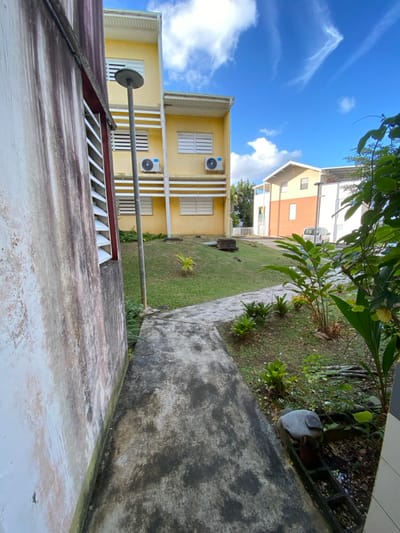 The scene depicts an outdoor residential or institutional area in Capesterre-Belle-Eau, France, under a clear blue sky with scattered white clouds. In the foreground, a stained and weathered light-colored concrete wall with a louvered window opening runs vertically along the left edge. A dark, stained concrete pathway, approximately 1.5 meters wide, extends from the immediate foreground towards the center-right, curving gently to the right. To the right of the pathway, a manicured grass lawn is interspersed with landscaped beds containing various green plants and foliage.
In the midground, a prominent three-story building, painted light yellow, features multiple horizontal louvered windows and two white external air conditioning units mounted on the second floor. A tall, slender grey street light fixture with a circular top stands on the grass between the concrete wall and the yellow building. Behind the yellow building, another building with a light-colored upper section and an orange-brown lower section is visible. A white vehicle, possibly a compact car or van, is partially visible parked on a paved surface beyond the buildings in the distance. The overall environment suggests a tropical or subtropical climate, characterized by the vegetation and architectural style. No individuals are present.