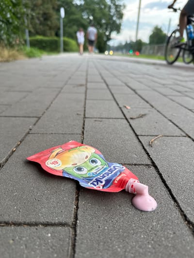 On a paved walkway in Sosnowiec, Poland, a fallen pouch of yogurt lies on its side, with pink yogurt spilled out of its open spout. The pouch features a cartoon character and Polish text. In the background, two individuals walk away from the camera, and a person on a bicycle rides past. The scene is set during the daytime, with trees and a street light visible.