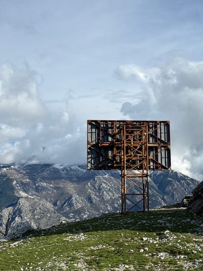 The image displays an outdoor mountainous landscape in Bogdašići, Montenegro. A large, heavily rusted metal structure, consistent with the rear framework of a billboard, stands centrally in the midground. This structure exhibits extensive corrosion and is composed of a complex lattice of horizontal, vertical, and diagonal support beams. The foreground consists of a grassy slope sparsely dotted with numerous light-colored rocks. In the background, rugged mountains rise, with their upper sections partially snow-covered and obscured by white and gray clouds. The sky above is overcast with varying cloud densities. No persons or discernible activity are present.