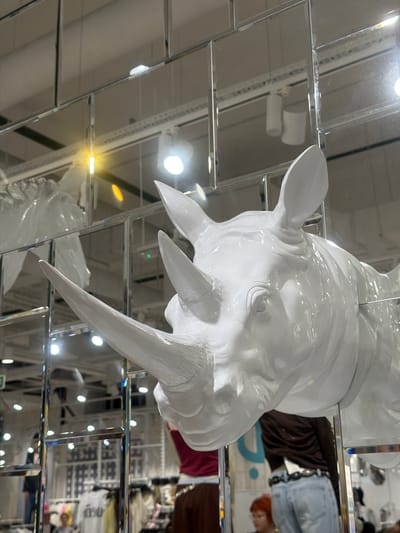 A low-angle shot shows a white, glossy sculpture of a rhino's head hanging from a mirrored ceiling. The reflections in the ceiling show a clothing store with visible garments and shoppers. The location is indeterminate and the description of being in Kuleshovka, Russia, is unconfirmed by visual evidence.