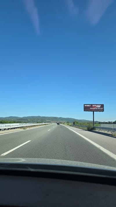 The image depicts a highway in Mursalevo, Bulgaria, under a clear blue sky. A roadside billboard displaying the "WINBET SCRATCH" advertisement stands along the side of the road. The asphalt highway has clearly marked white lines. The scene is captured from inside a vehicle with a view out onto the road and countryside.